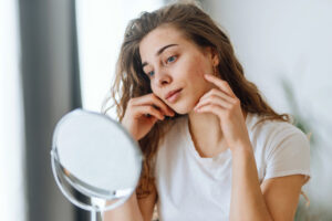 Young woman looking closely at the dark spots on her face in the mirror, in need of the VI Peel.